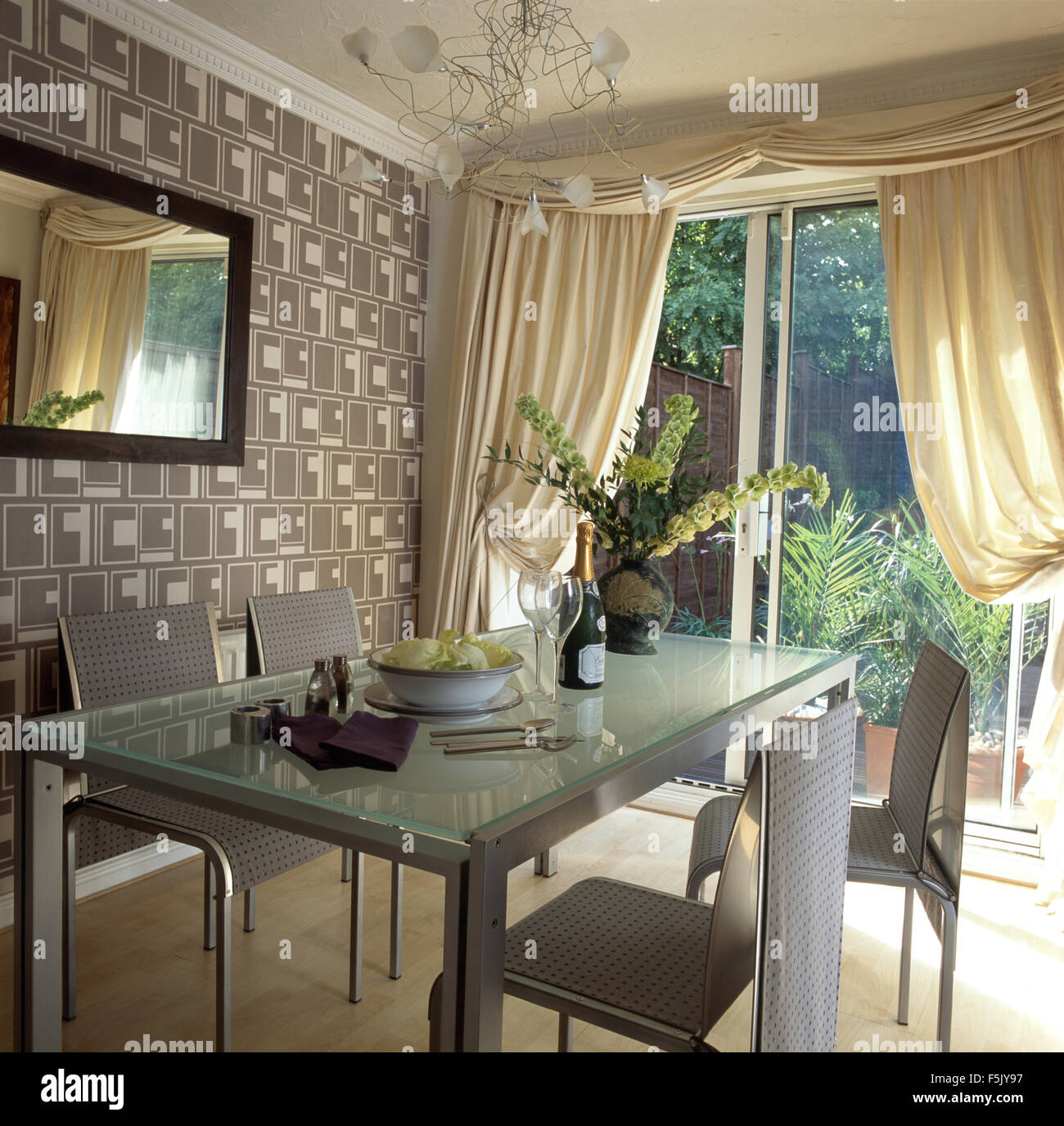 Des chaises en métal et verre table de salle à manger avec abstrait gris papier peint à motifs et des rideaux crème sur les portes en verre Banque D'Images
