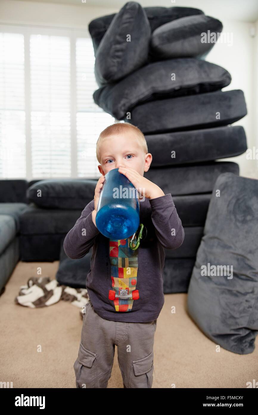 Boy drinking in living room, pile de coussins en arrière-plan Banque D'Images