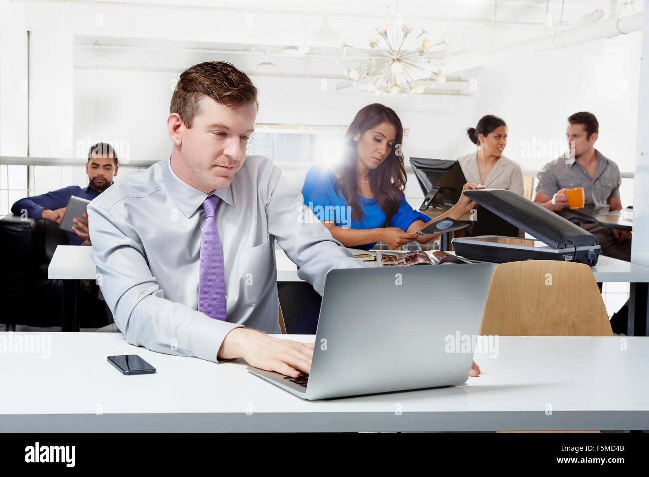 Mid adult businessman sitting at desk, à l'aide d'un ordinateur portable, des collègues qui travaillent en arrière-plan Banque D'Images