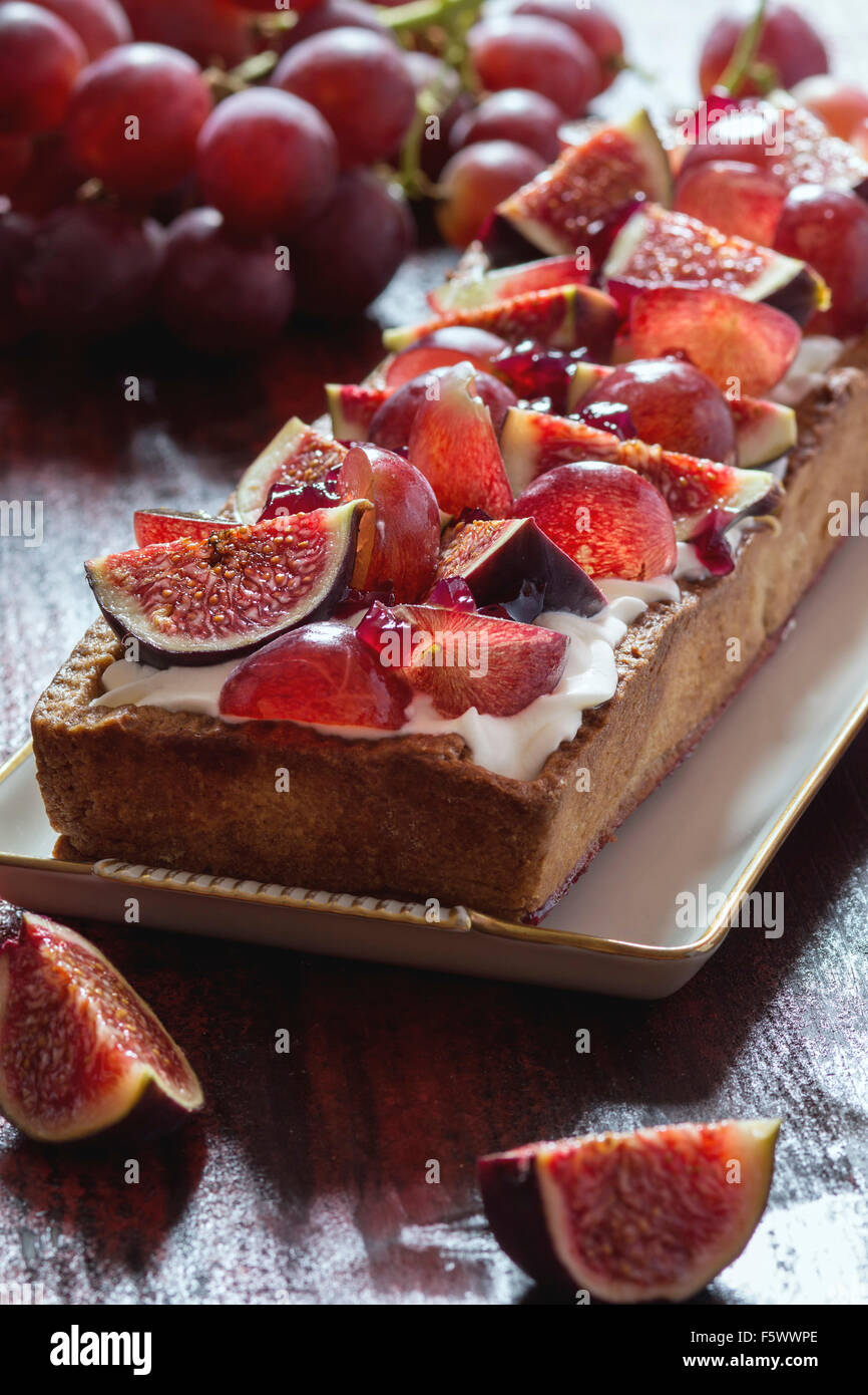 Tarte rectangulaire rouge avec des raisins, figues et à la crème fouettée dans la plaque en céramique blanc sur rouge table en bois. Rustique foncé styl Banque D'Images