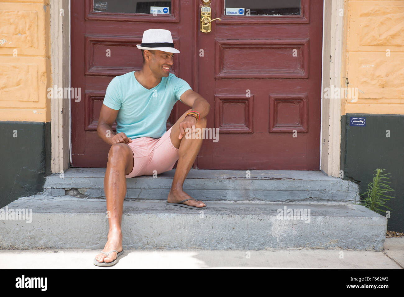 Un homme portant une ambigu t-shirt vert clair, rose short, lunettes et fedora sur une véranda à l'extérieur d'une maison avec Burnt Umber doubles portes. Banque D'Images