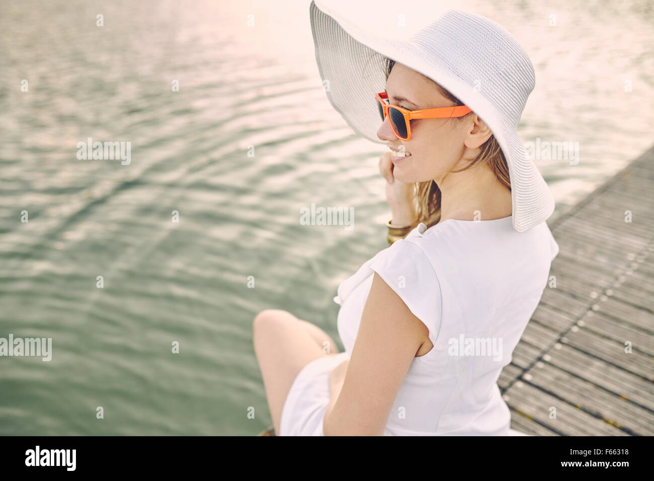 Portrait d'une femme assis sur un quai avec robe blanche et chapeau et verre orange Banque D'Images