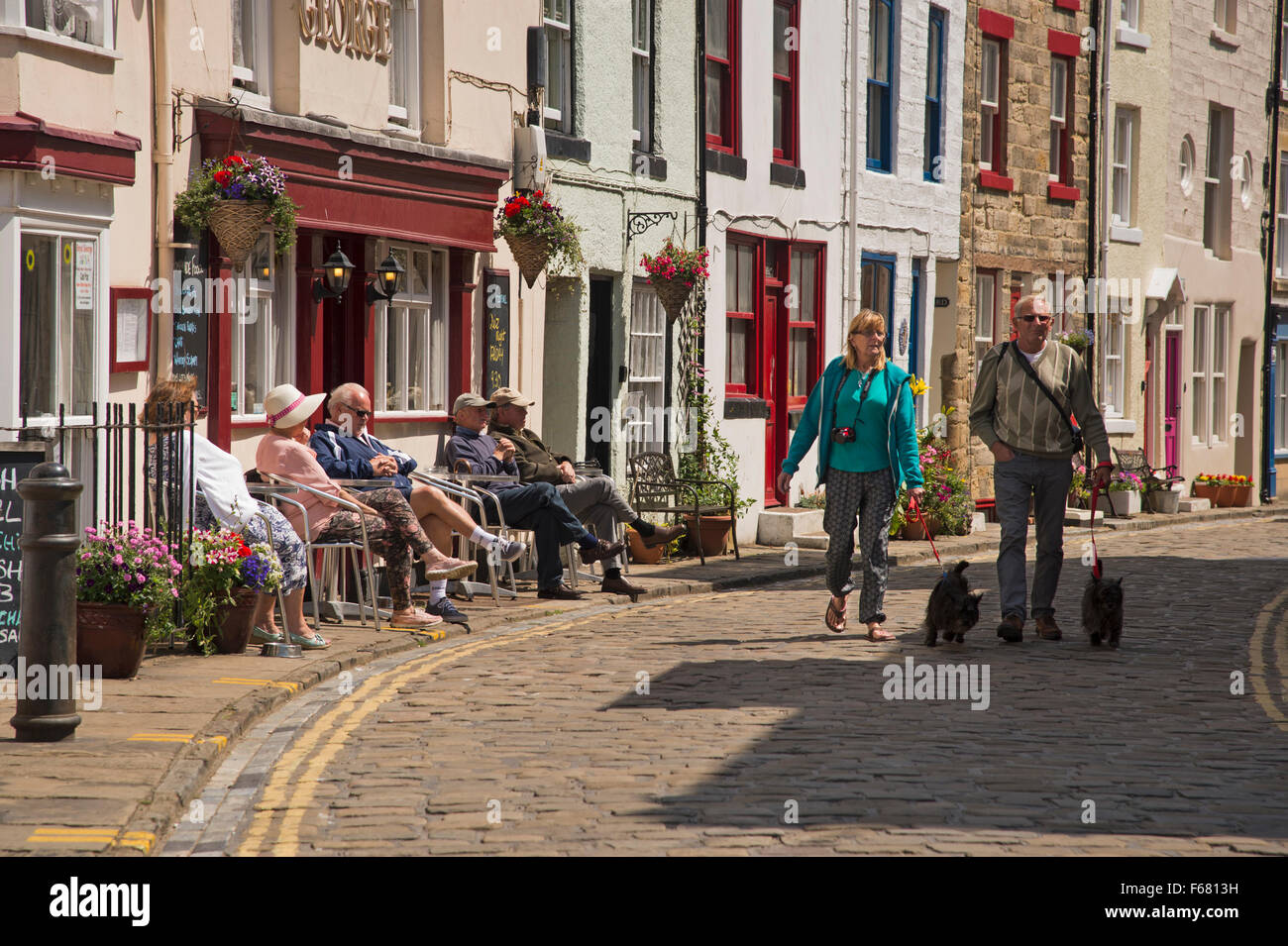 Les gens se détendre, assis à l'extérieur d'un pub ou marcher le long de la rue principale pavée ensoleillée - village de pêche traditionnel de Staithes, North Yorkshire, UK. Banque D'Images