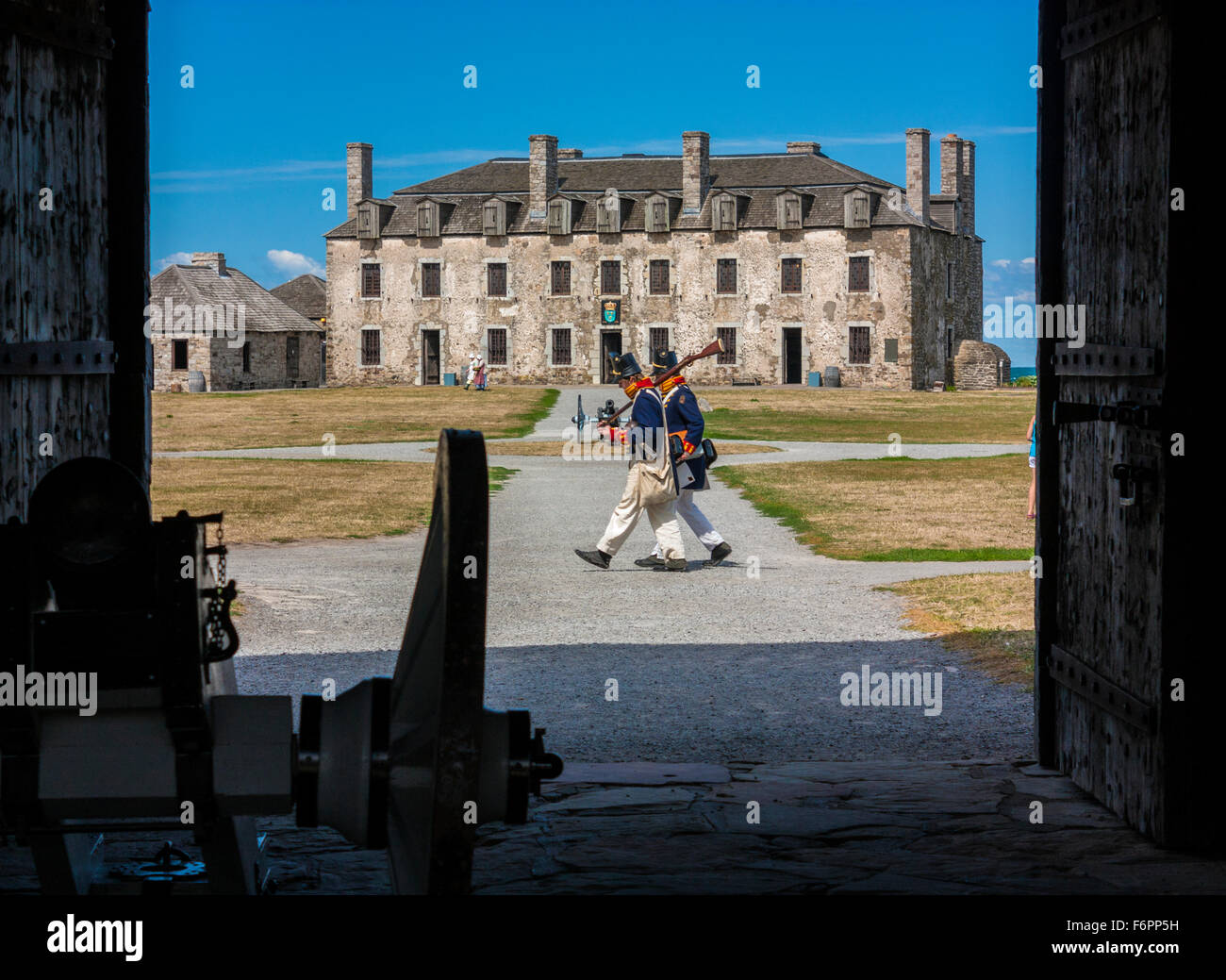 Deux soldats en uniformes d'époque traversent une cour de fort historique, avec de vieux bâtiments en pierre au loin encadrés par la porte d'entrée. Banque D'Images