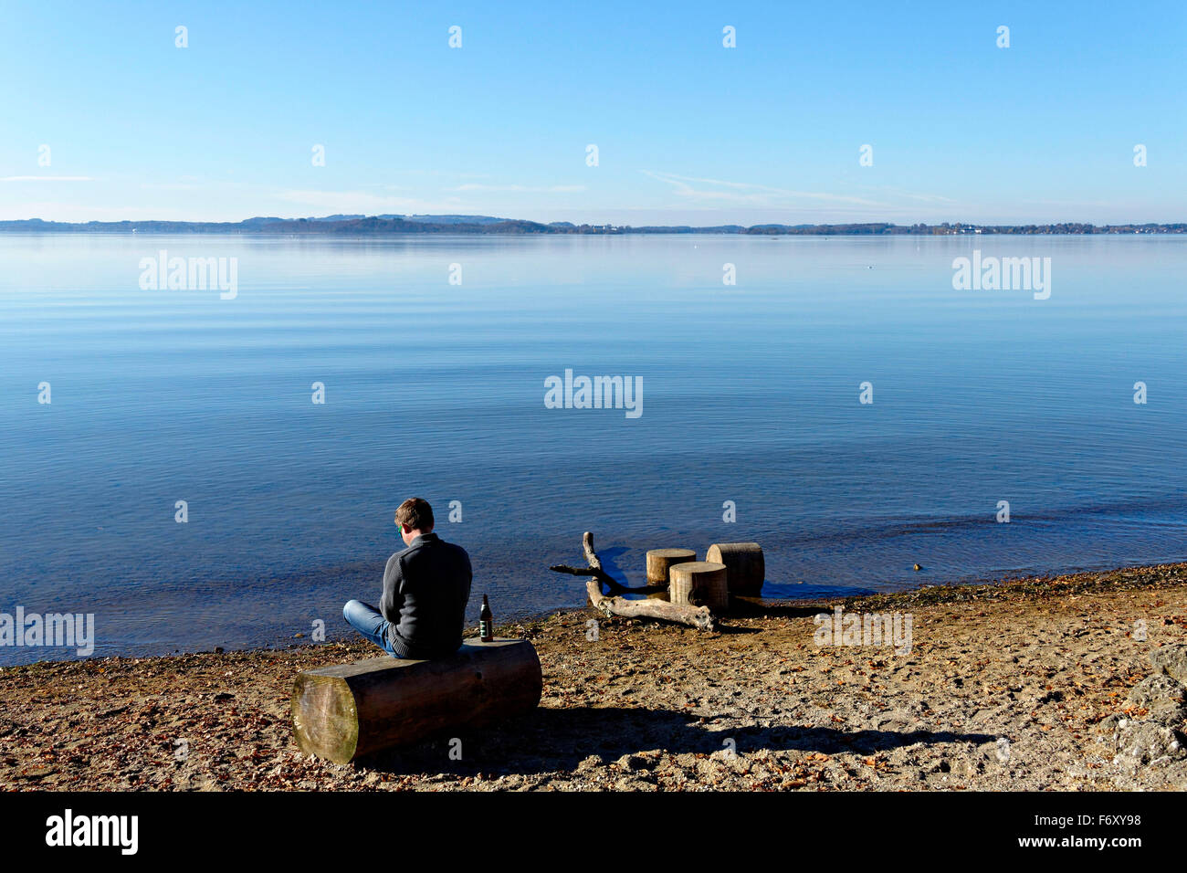 Homme assis sur un journal en bois avec une bière au bord d'un lac, Feldwieser Bay, Chiemsee, Upper Bavaria, Germany, Europe. Banque D'Images