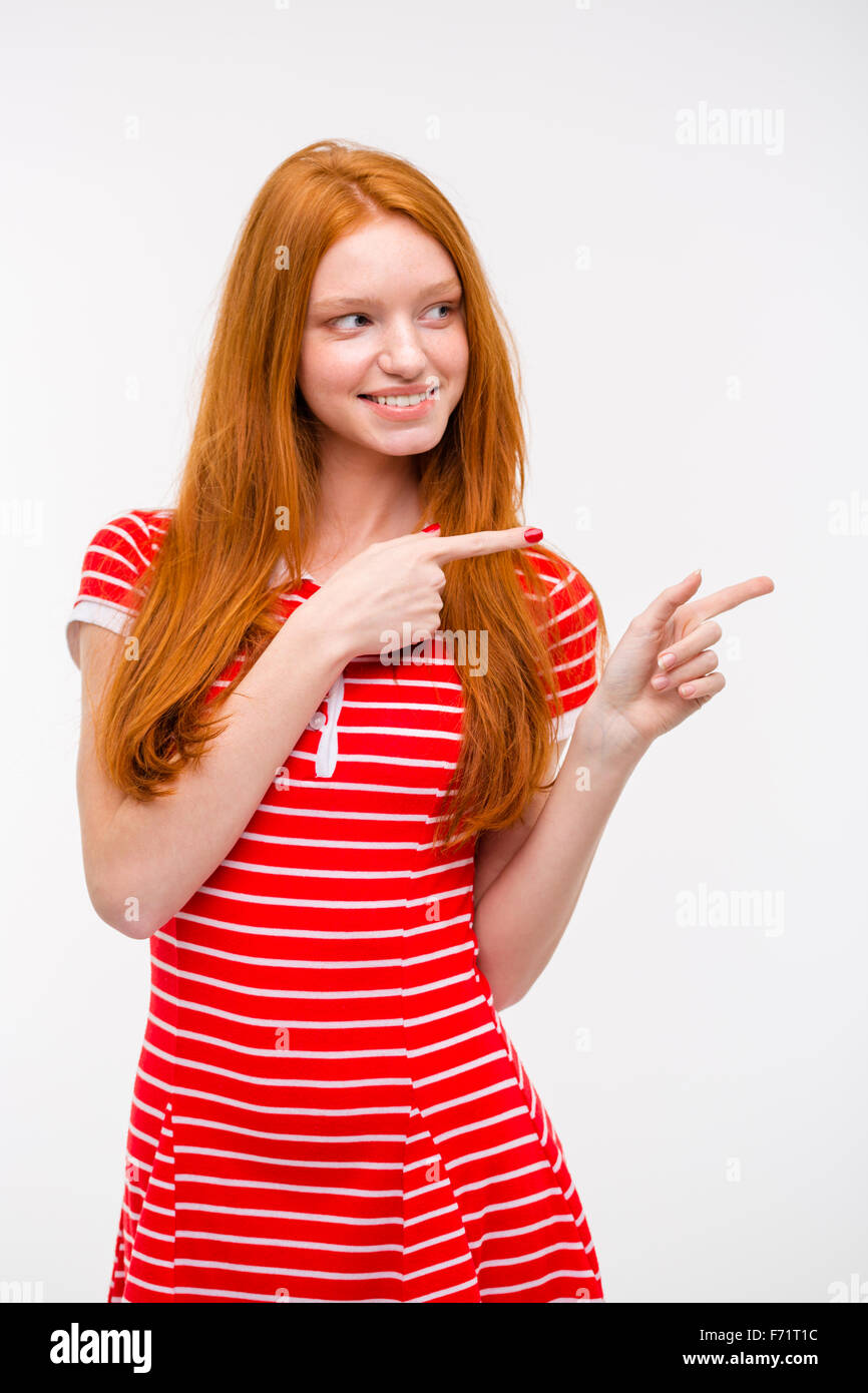 Cheerful belle jeune femme rousse en robe rayée rouge dirigés à l'écart tout en standing against white background Banque D'Images