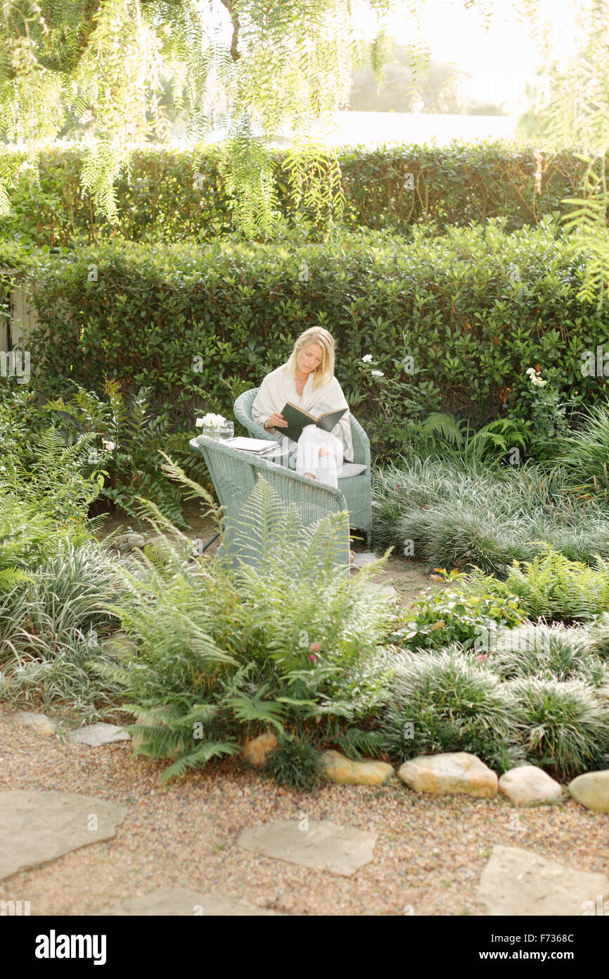 Femme blonde assise dans un fauteuil en osier dans un jardin, la lecture. Banque D'Images Femme blonde assise dans un fauteuil en osier dans un jardin, la lecture. Banque D'Images