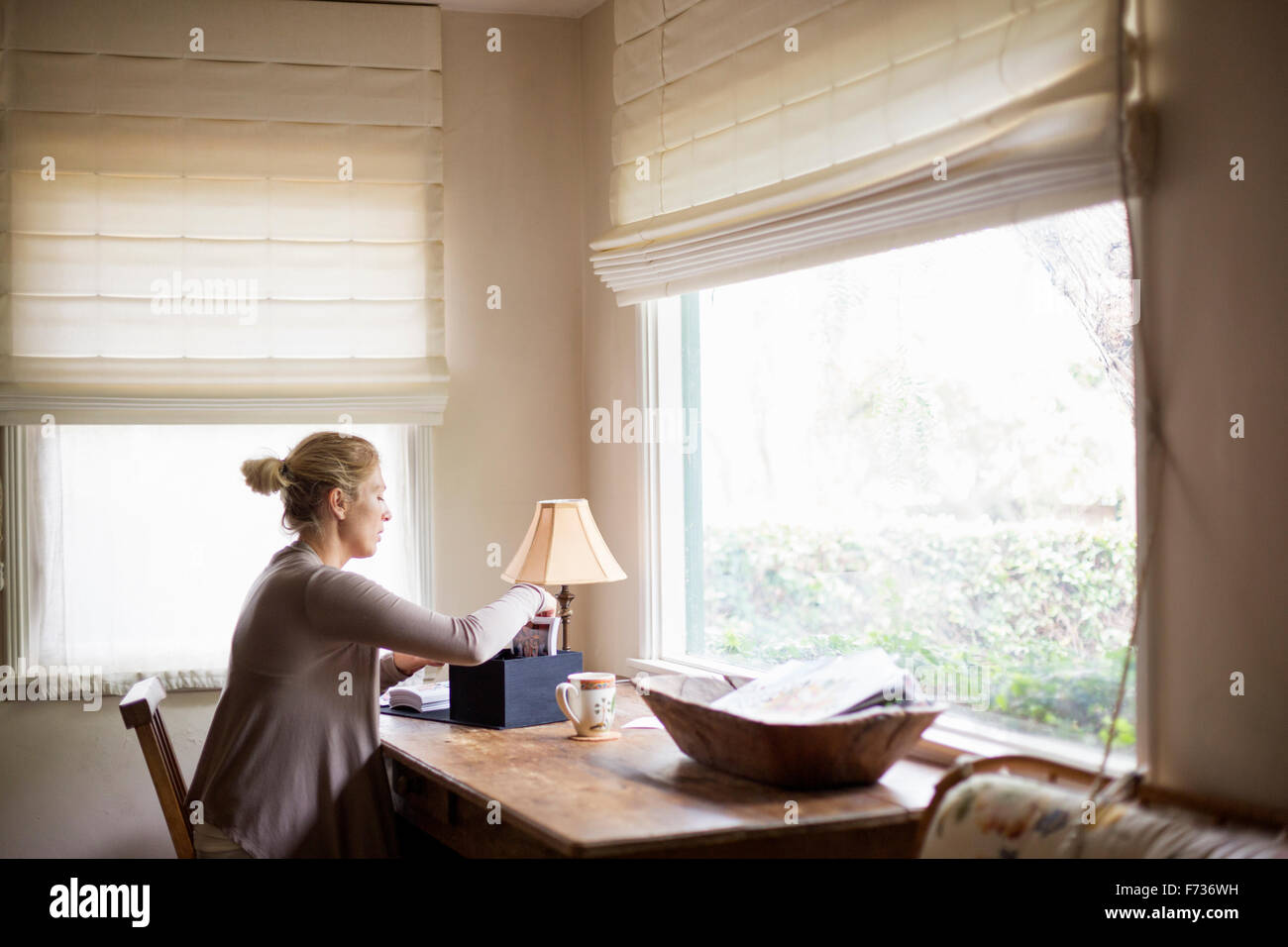 Femme blonde assis à un bureau par une fenêtre, le tri d'une boîte avec des photos. Banque D'Images