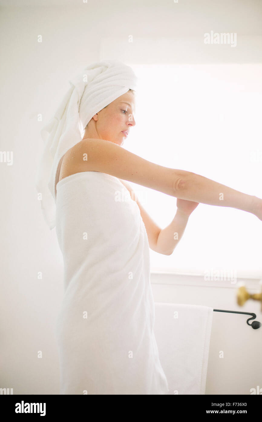 Femme enveloppée dans une serviette blanche debout dans une salle de bain, l'application de lotion sur son bras. Banque D'Images