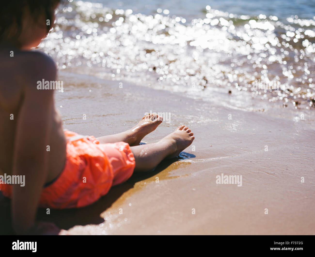 Un garçon assis sur le sable au bord de l'eau regardant les vagues à la plage. Banque D'Images