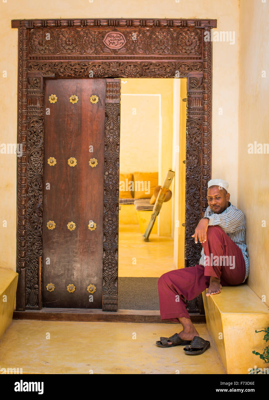 LAMU, Kenya, Afrique. Un homme en robe arabe traditionnelle se trouve sur un becnh à l'extérieur une ancienne porte en bois sculpté. Banque D'Images