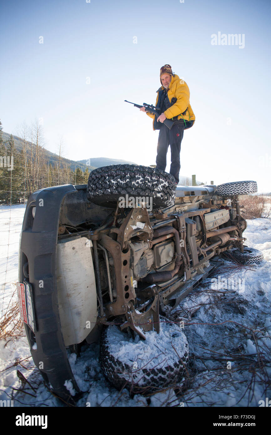 Un chasseur tient sa rifel en étant debout sur son camion retourné après un accident. Banque D'Images