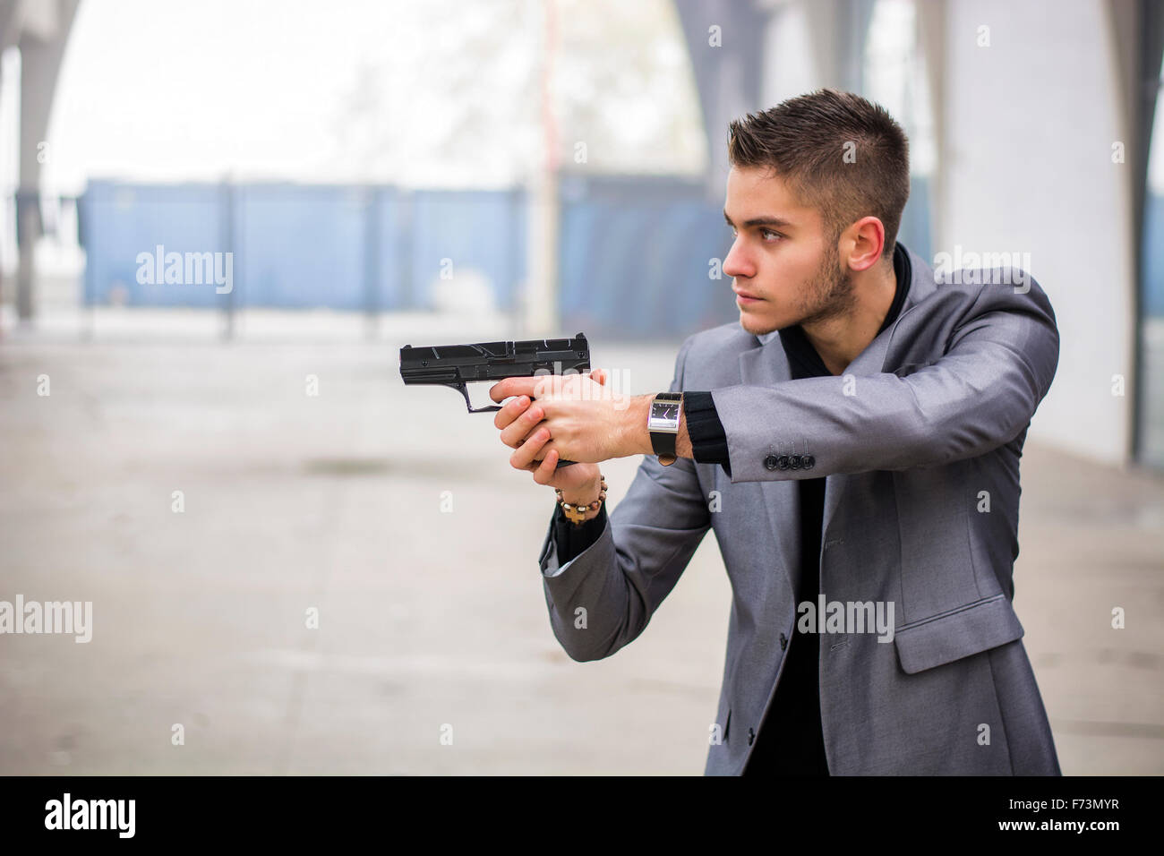 Bien habillé beau jeune détective ou policier ou gangster debout dans un environnement urbain visant une arme à feu sur la gauche Banque D'Images