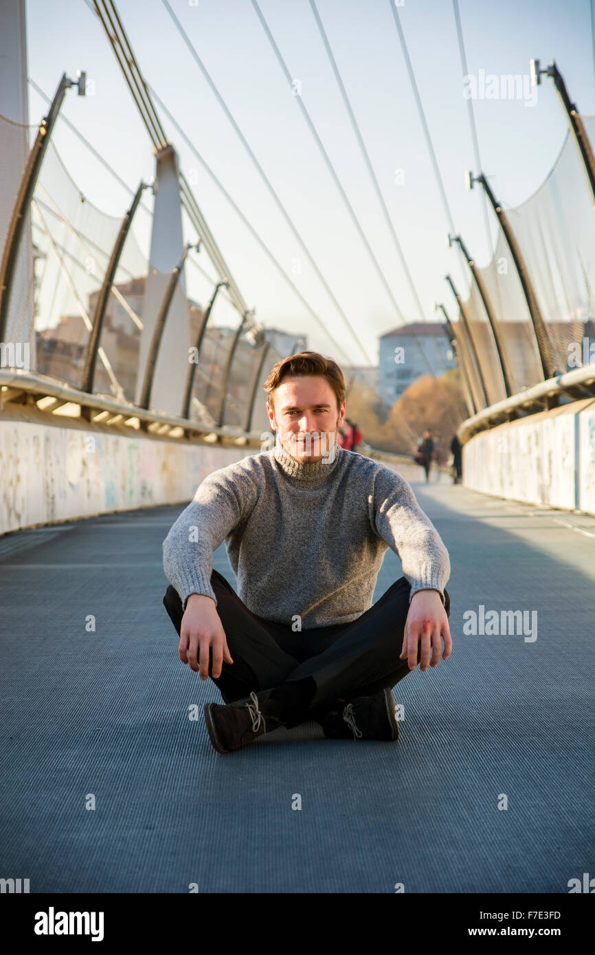 Beau jeune homme assis à l'extérieur dans l'environnement urbain, portant des vêtements d'hiver Banque D'Images