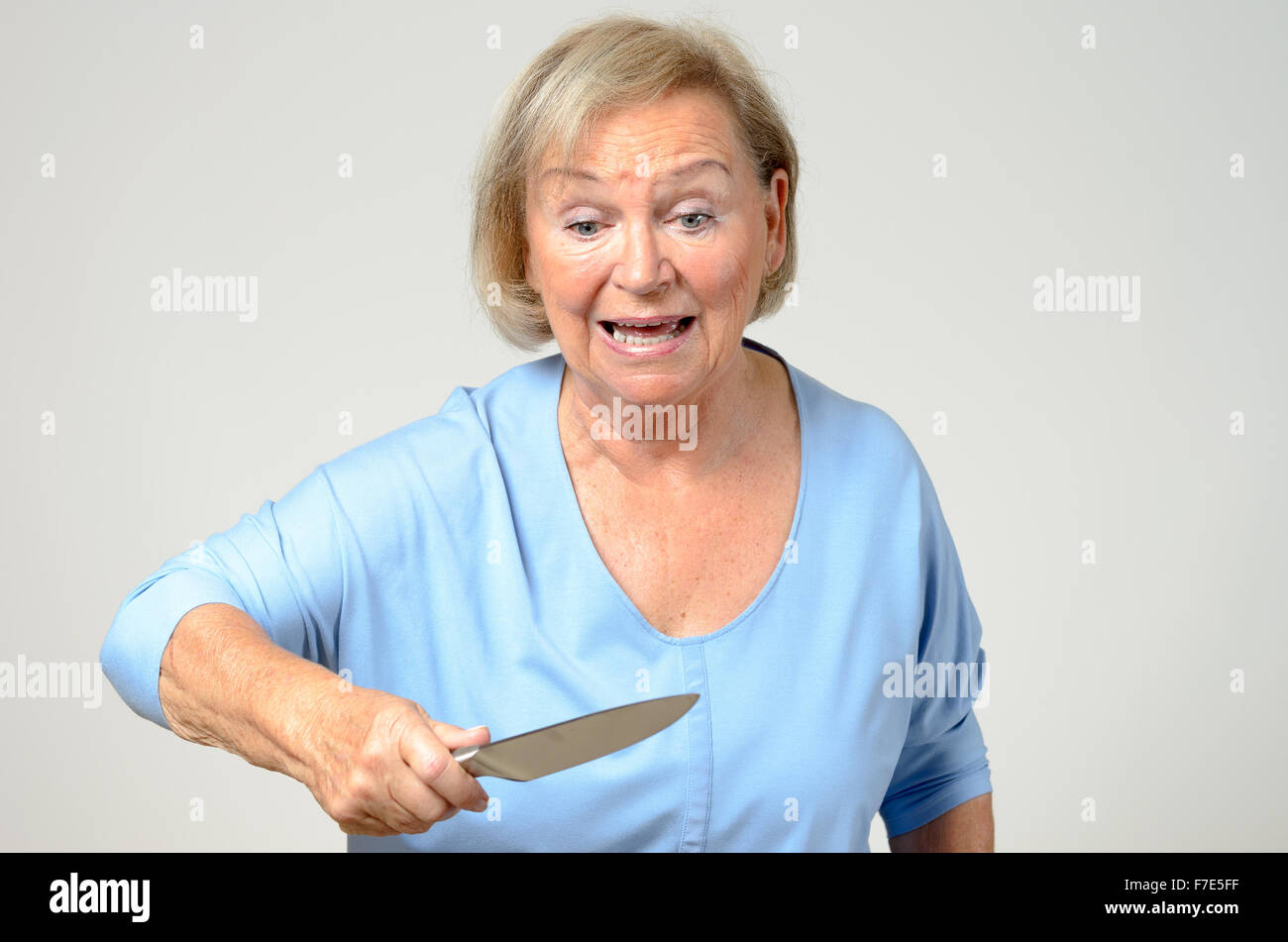 Femme âgée émotionnelle brandissant un couteau de cuisine dans sa main avec un regard perplexe de l'horreur et de choc, plus de Gray Banque D'Images
