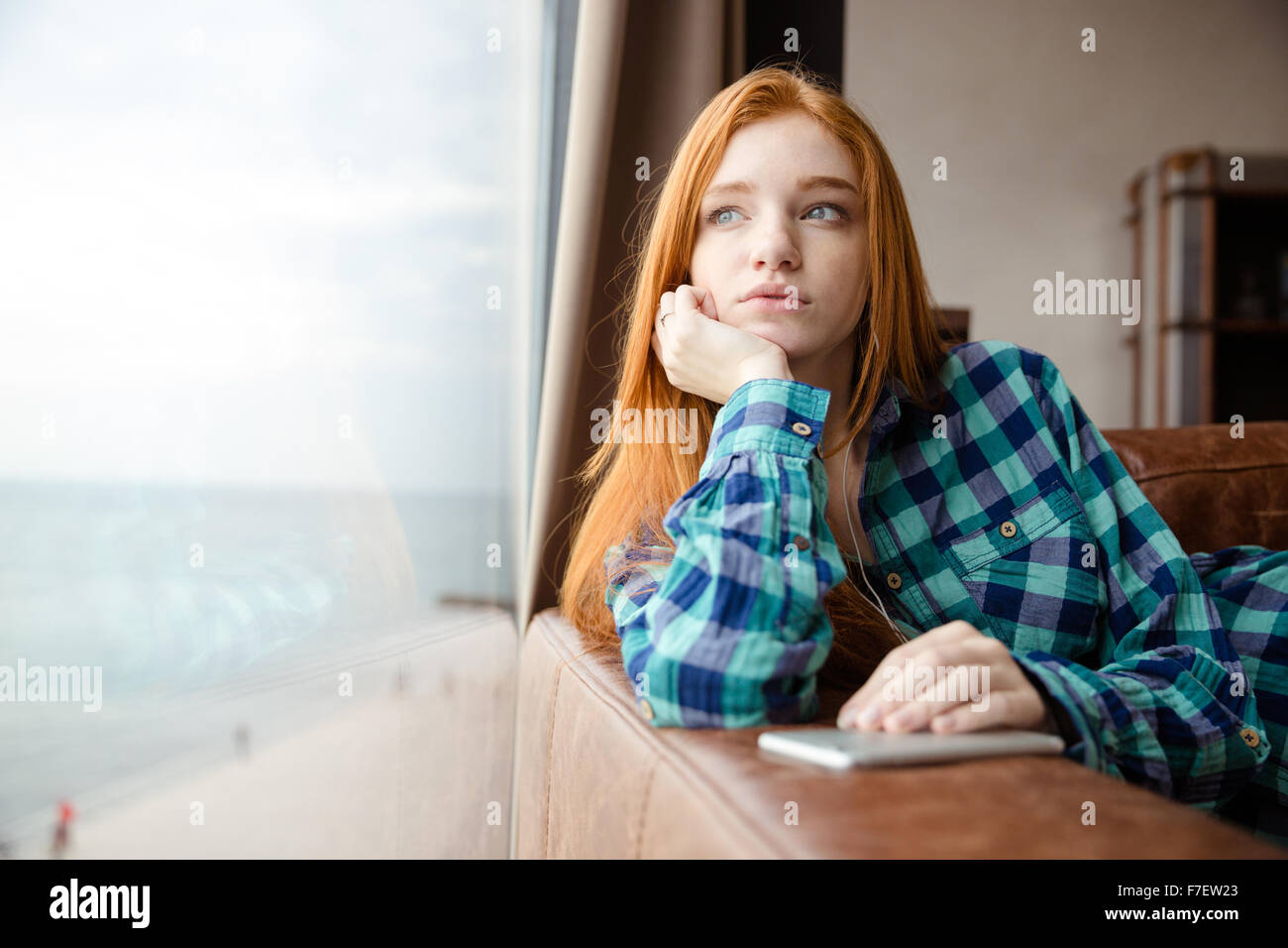 Réfléchis rêve belle jolie fille avec de longs cheveux rouge chemise à carreaux en regardant par la fenêtre et écouter de la musique Banque D'Images