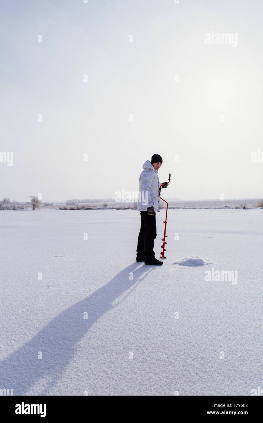 Pêcheur d'hiver avec broche à glace faire trou dans la glace sur un lac gelé Banque D'Images