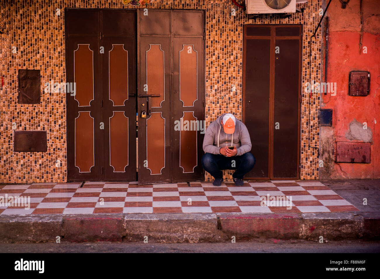 Homme marocain en utilisant son téléphone mobile tout en s'accroupissant au soleil dans une rue de Marrakech Banque D'Images