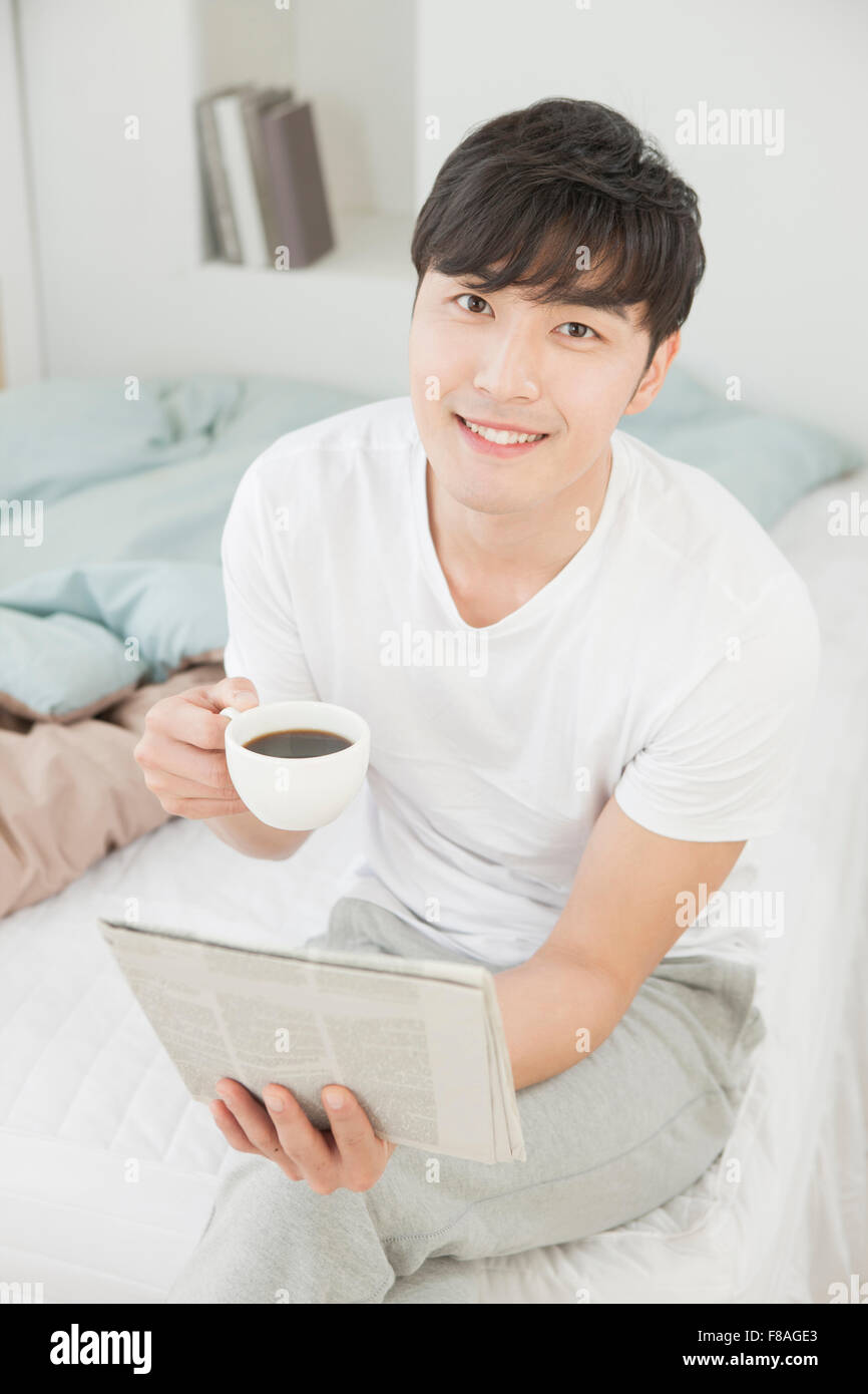 High angle de l'homme assis sur le lit avec les jambes croisées tenant une tasse de café et regarder le journal de l'avant Banque D'Images