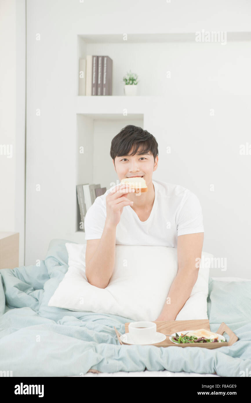 Homme assis dans le lit de mordre une tranche de pain avec le plateau du petit déjeuner Banque D'Images