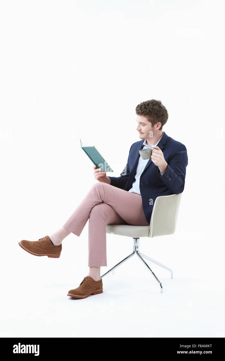 Businessman sitting on chair les jambes croisées, la tenue et la lecture d'un livre avec un verre à la main Banque D'Images