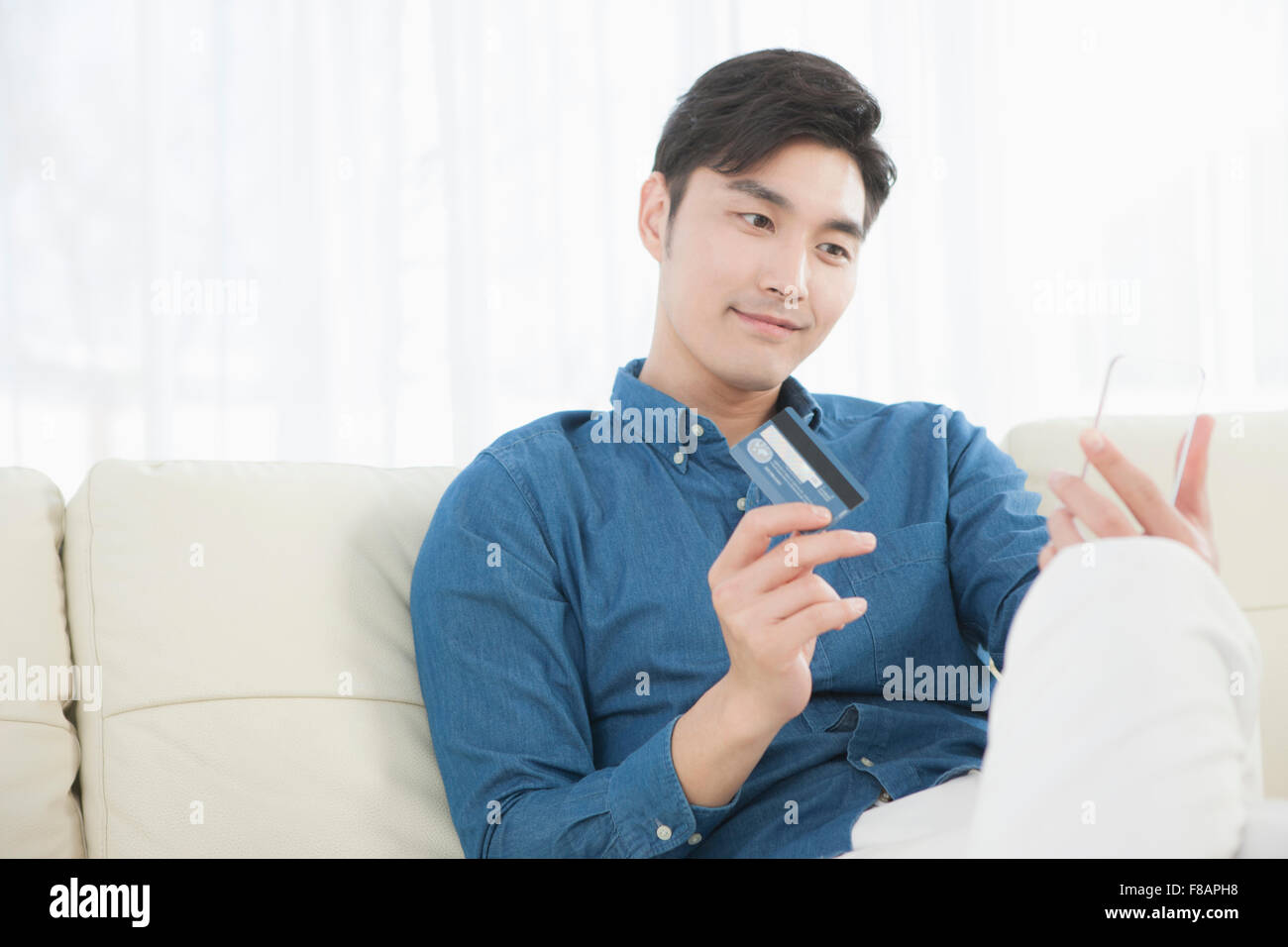 Jeune homme tenant une carte de crédit et un smartphone transparent sitting on sofa Banque D'Images