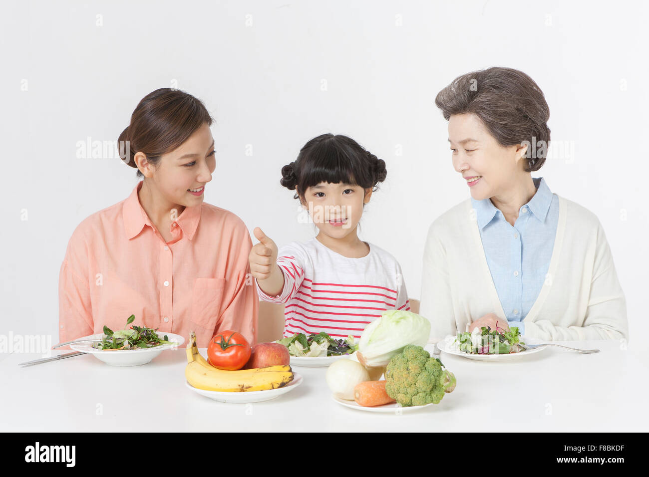 Jeune fille avec des geste de la main, sa mère, et la grand-mère assis à table avec des assiettes à salade et variété de légumes Banque D'Images