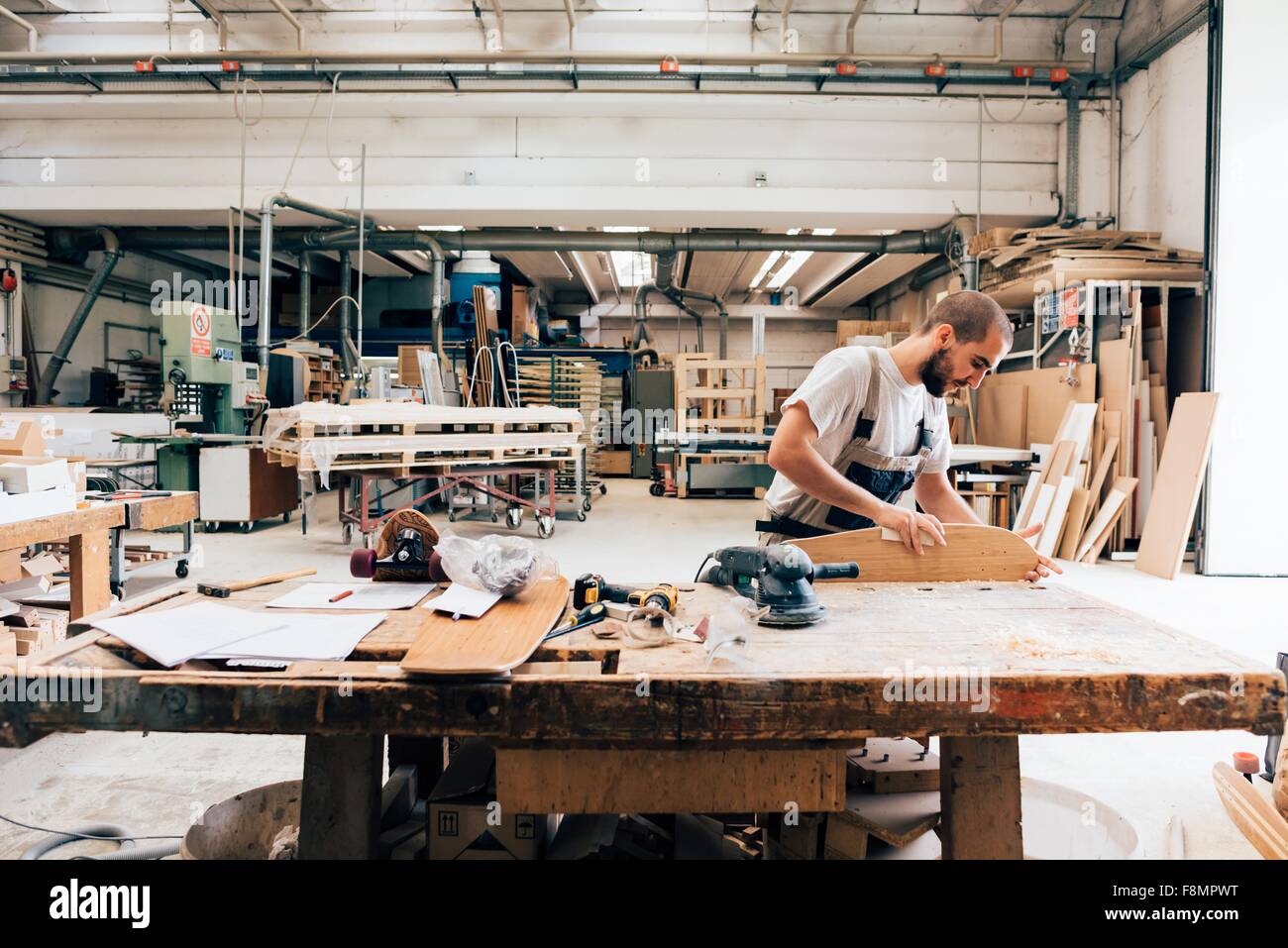 Jeune homme en atelier de menuiserie à la recherche vers le bas de la planche à roulettes de ponçage Banque D'Images