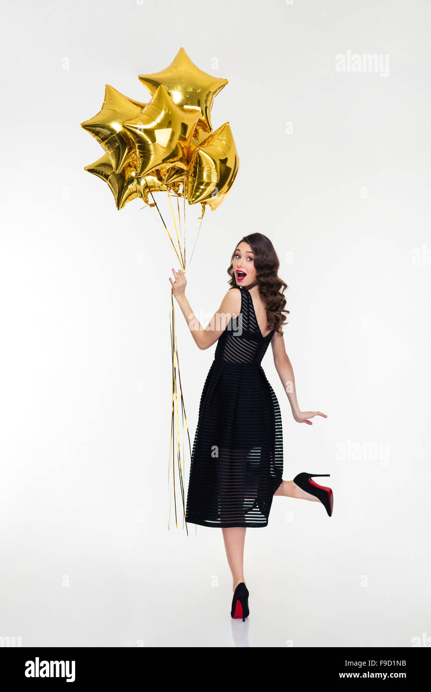 Jolie femme heureux ludique avec retro hairstyle classique en robe noire et des chaussures à l'arrière et tenant des ballons d'or Banque D'Images