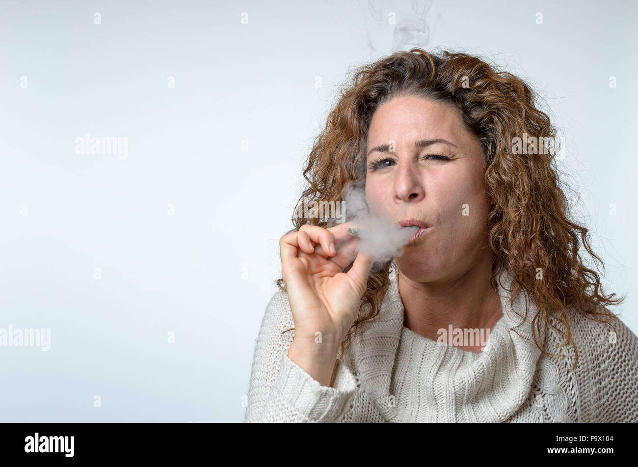 Belle jeune femme que quelques bouffées sur une cigarette avec un oeil fermé dans la jouissance sur un arrière-plan gris studio Banque D'Images