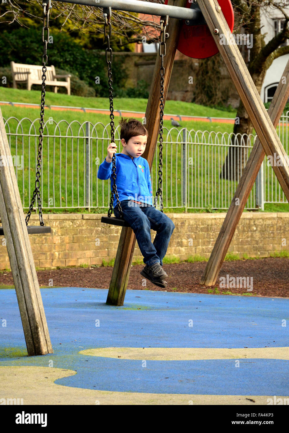 Un garçon d'une veste bleue est assis sur une balançoire dans un aire de jeux pour enfants. Banque D'Images