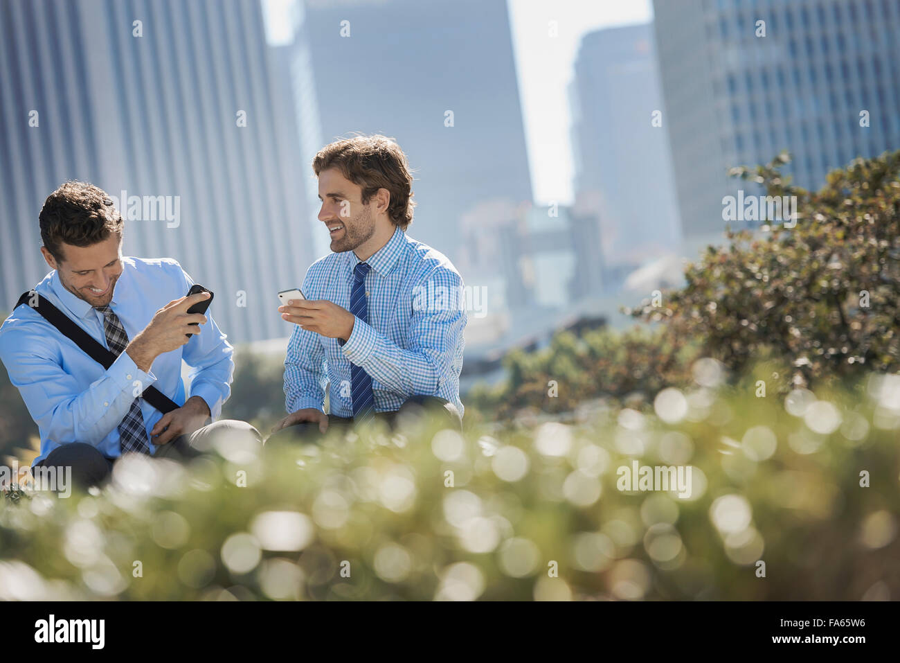 Deux hommes en chemises et cravates dans un parc dans la ville tenant leurs téléphones intelligents. Banque D'Images