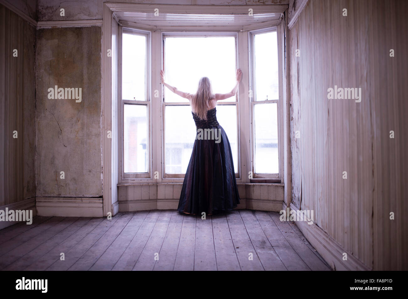 Une jeune femme aux cheveux blonds mince portant une longue robe de bal robe seul dans une chambre à l'abandon de la réduction des capacités à la recherche permanente d'une grande baie vitrée UK Banque D'Images