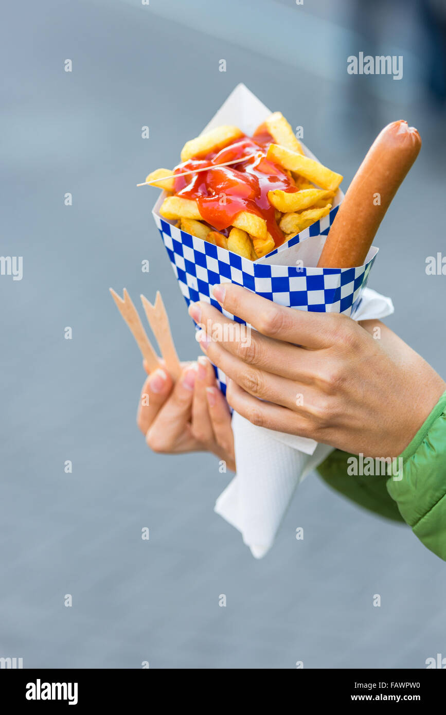Les mains tenant le cône de papier avec une saucisse chaude et de frites avec du ketchup Banque D'Images