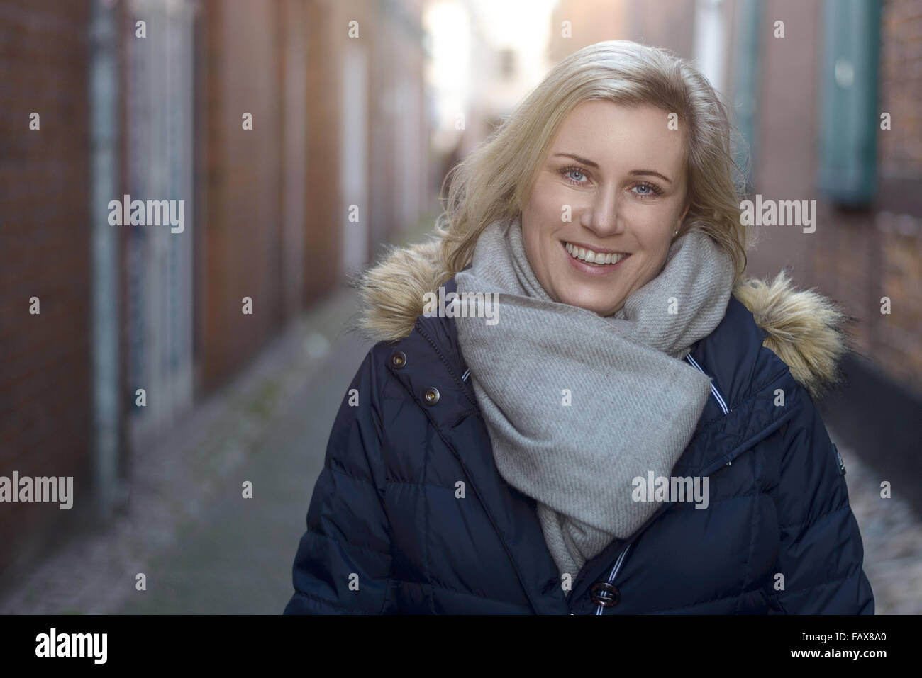 Jolie blonde amical femme portant une écharpe d'hiver élégant debout dans une ruelle de la ville souriant joyeusement à l'appareil photo Banque D'Images