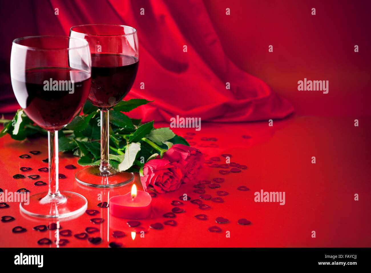 Une vie toujours romantique avec deux verres de vin rouge, une bougie en forme de coeur et deux roses, avec un fond de satin. Banque D'Images