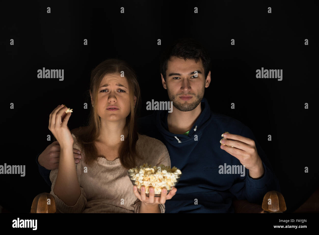 Jeune couple, homme et femme, assise dans la pièce sombre à l'avant de regarder la télévision et film eating popcorn, montrant les émotions Banque D'Images