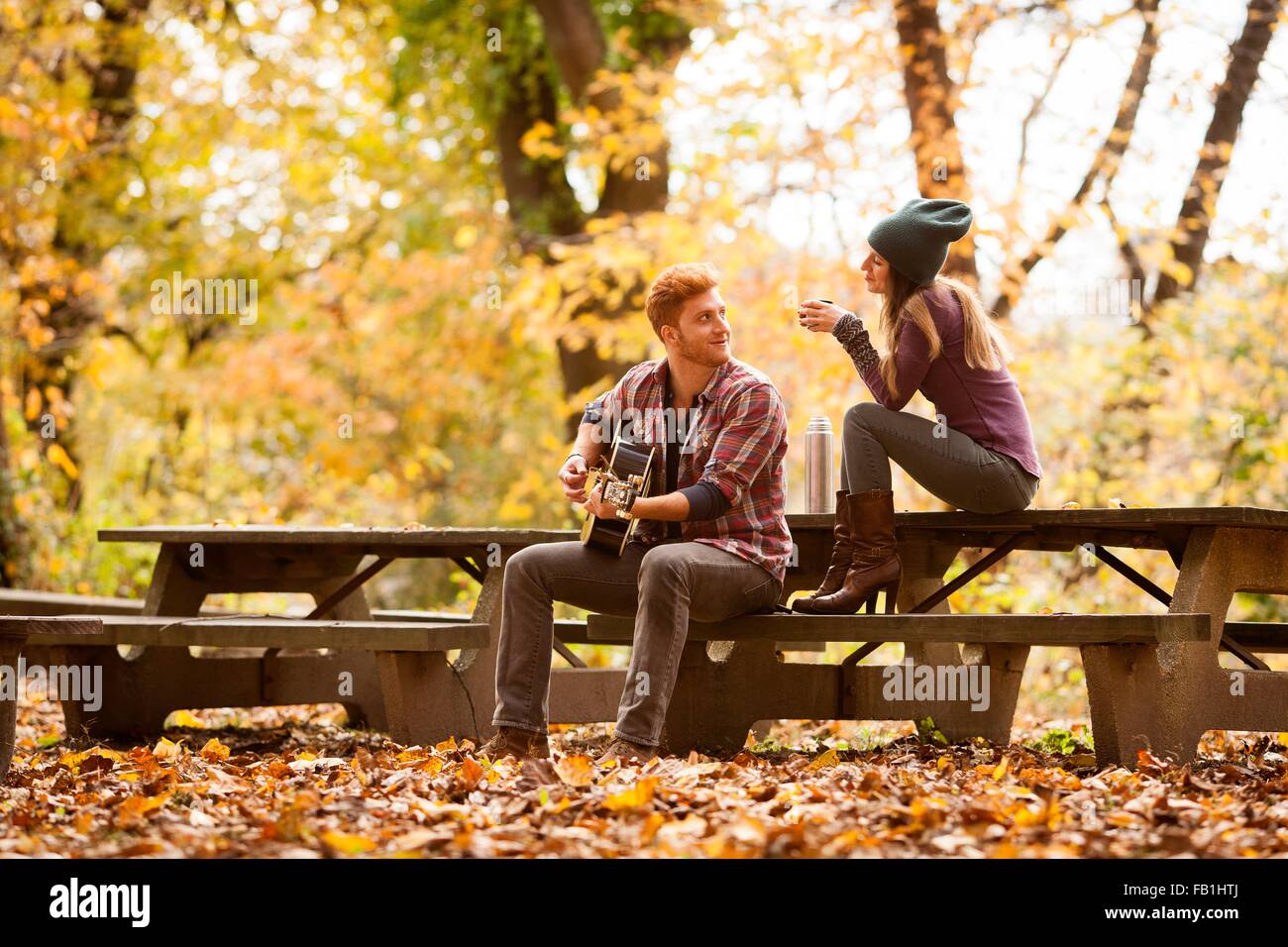 Jeune couple qui joue de la guitare sur des bancs de pique-nique en forêt d'automne Banque D'Images