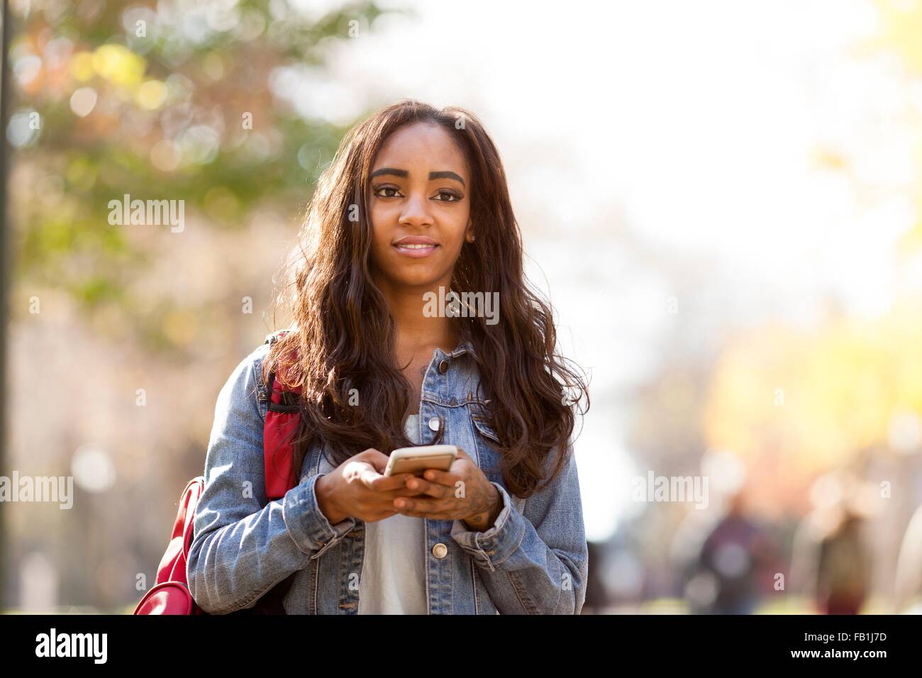 Jeune femme aux longs cheveux bruns portant veste denim holding smartphone à l'écart smiling Banque D'Images