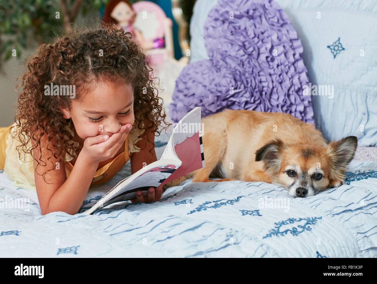 Girl and dog lying on bed reading book, main sur la bouche de rire Banque D'Images