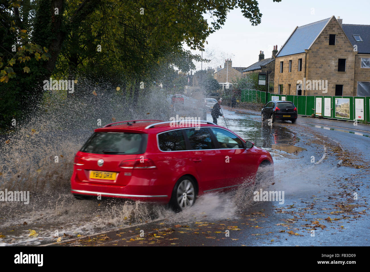 Après de fortes pluies, voiture conduit par grande flaque sur la route inondée, pulvériser de l'eau qu'il arrondit bend dans village. Burley dans Wharfedale, FR, UK. Banque D'Images