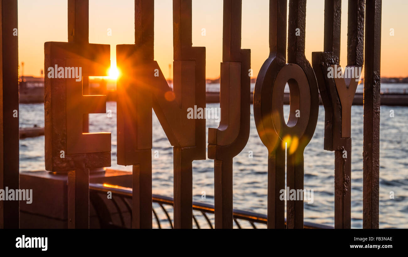 Le mot 'profiter' encadré en fer forgé au World Financial Plaza avec un coucher de soleil sur la rivière Hudson derrière Banque D'Images