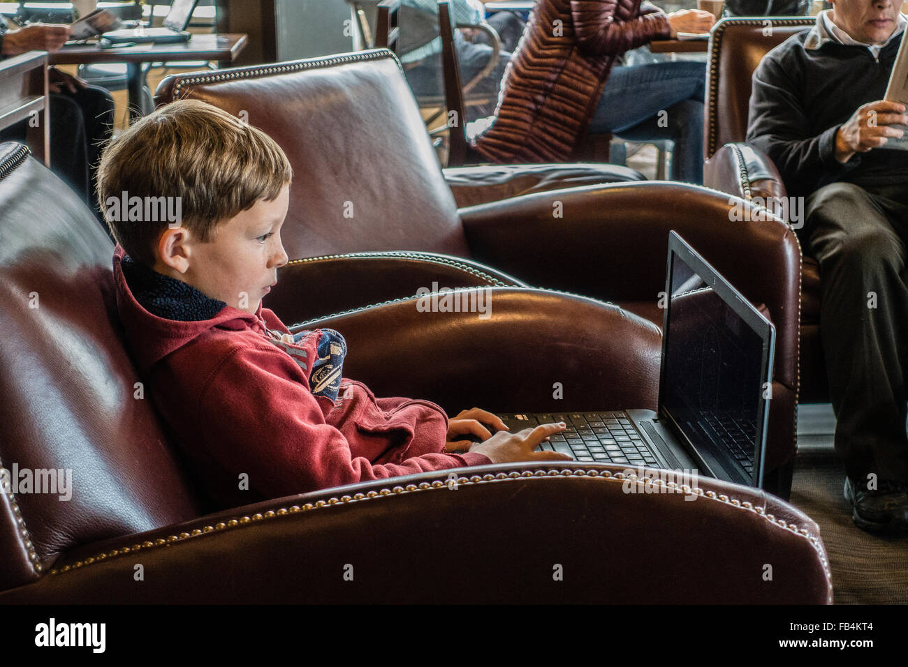 Un jeune garçon technophiles, de 10 à 12 ans, est assis dans un café avec un gros ordinateur portable sur ses genoux comme il se concentre sur elle. Banque D'Images