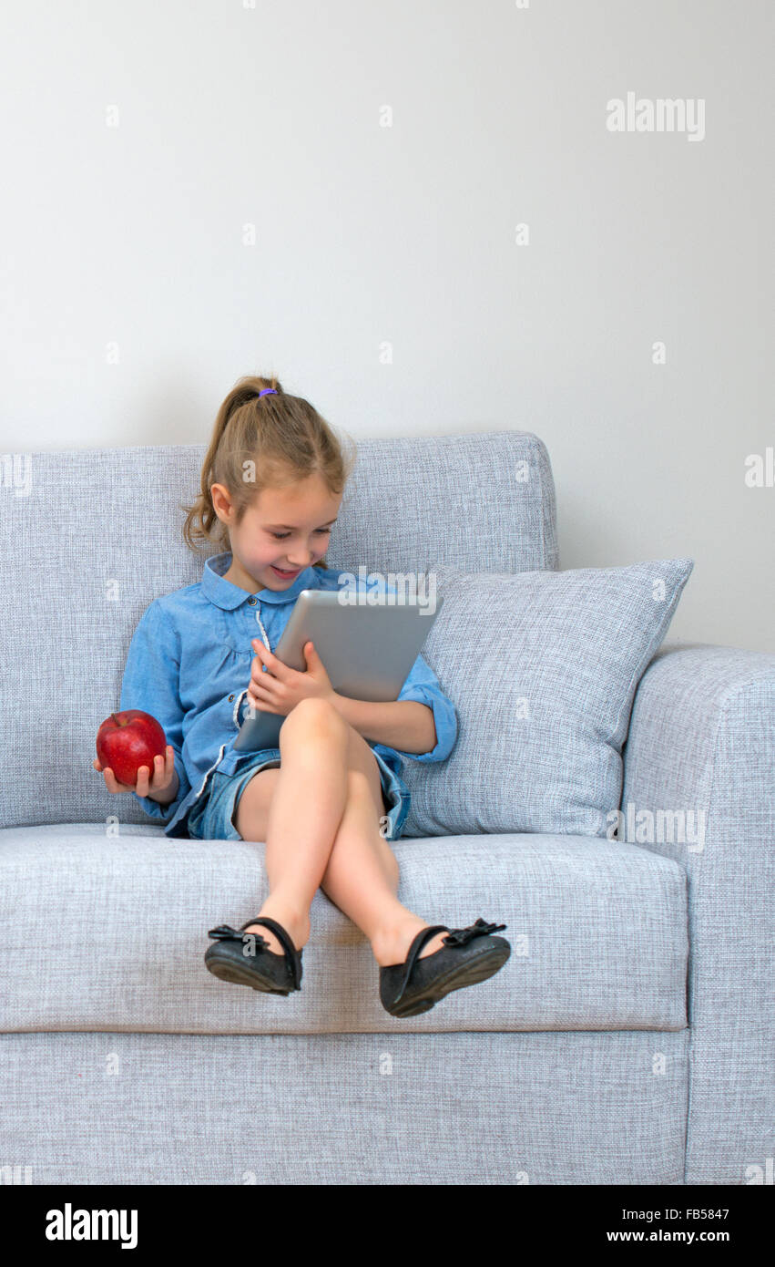 Petite fille assise sur le canapé et à l'aide de tablet pc. Banque D'Images