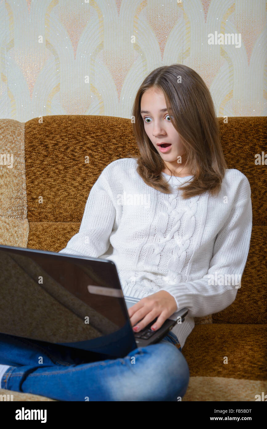 Une fille dans un chandail blanc et un jean assis sur le canapé avec votre ordinateur portable Banque D'Images