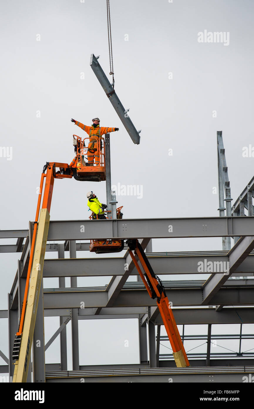 Les travaux de construction au Royaume-Uni : les hommes travaillent en hauteur sur des plates-formes 'cherry picker', boulonnage ensemble le cadre en acier d'un nouveau développement pour le supermarché Tesco et Marks & Spencer store sur un chantier à Aberystwyth, Pays de Galles, Royaume-Uni Banque D'Images