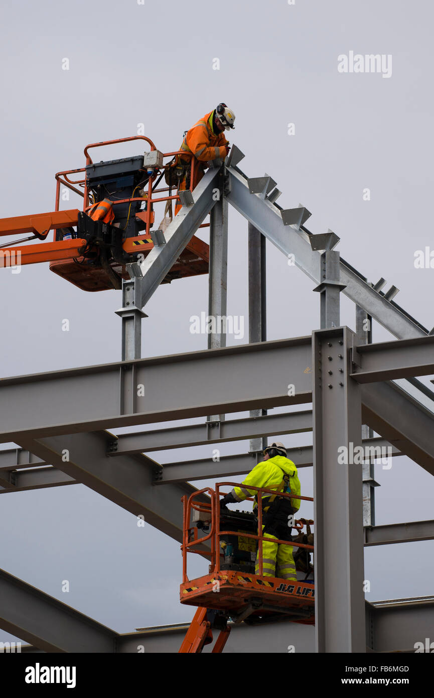 Les travaux de construction au Royaume-Uni : les hommes travaillent en hauteur sur des plates-formes 'cherry picker', boulonnage ensemble le cadre en acier d'un nouveau développement pour le supermarché Tesco et Marks & Spencer store sur un chantier à Aberystwyth, Pays de Galles, Royaume-Uni Banque D'Images