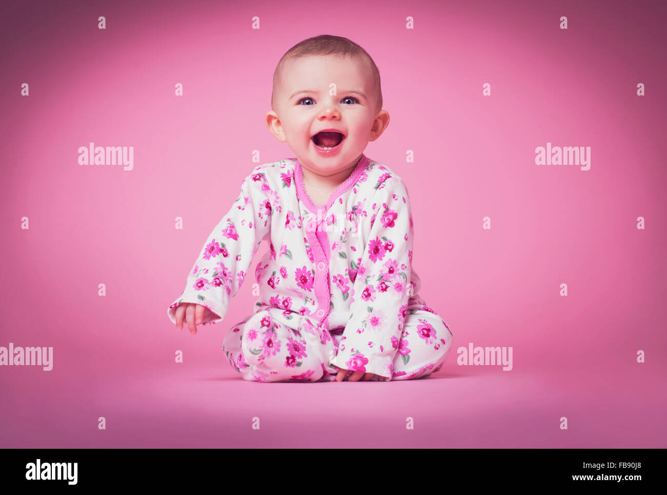 Cute smiling blue eyed baby girl in pink flower babygrow contre fond rose en studio. Rire. Banque D'Images