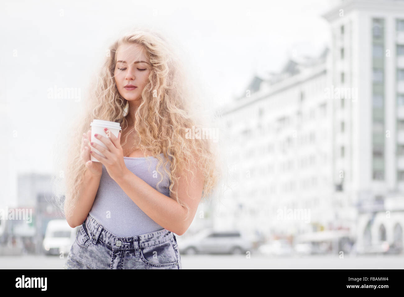 Heureux jeune femme avec de longs cheveux bouclés, tenant une tasse de café à emporter et souriant avec flirter devant une caméra contre les Banque D'Images