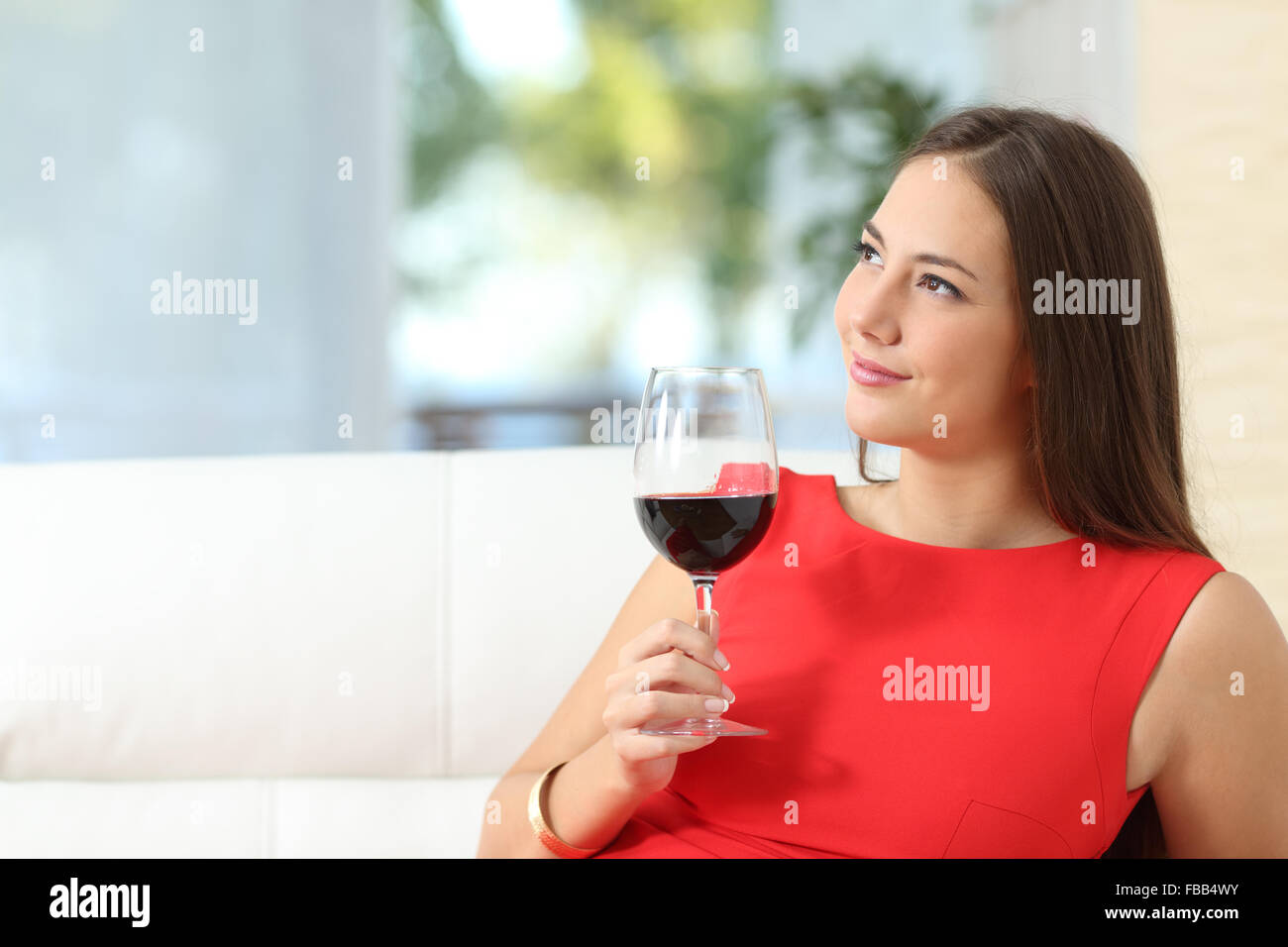 Pensive woman in red mode détendue avec une coupe de vin, assis sur un canapé à la maison Banque D'Images